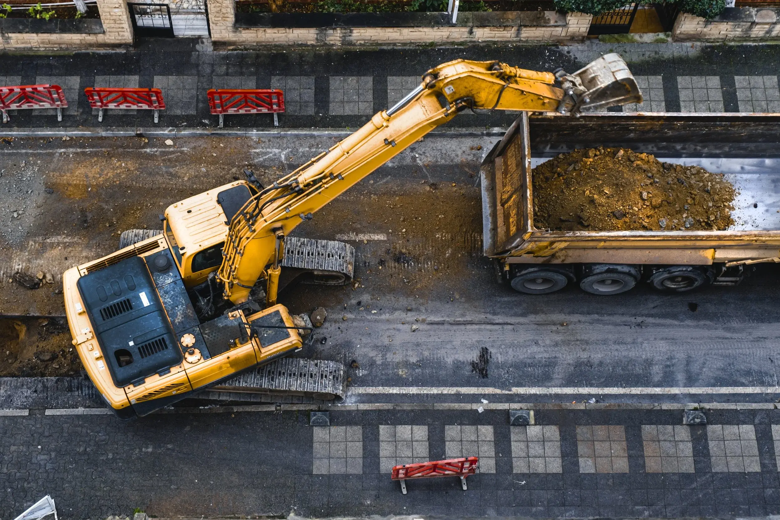 A yellow excavator on a road with red barrier fencing, loading dirt into a dump truck.